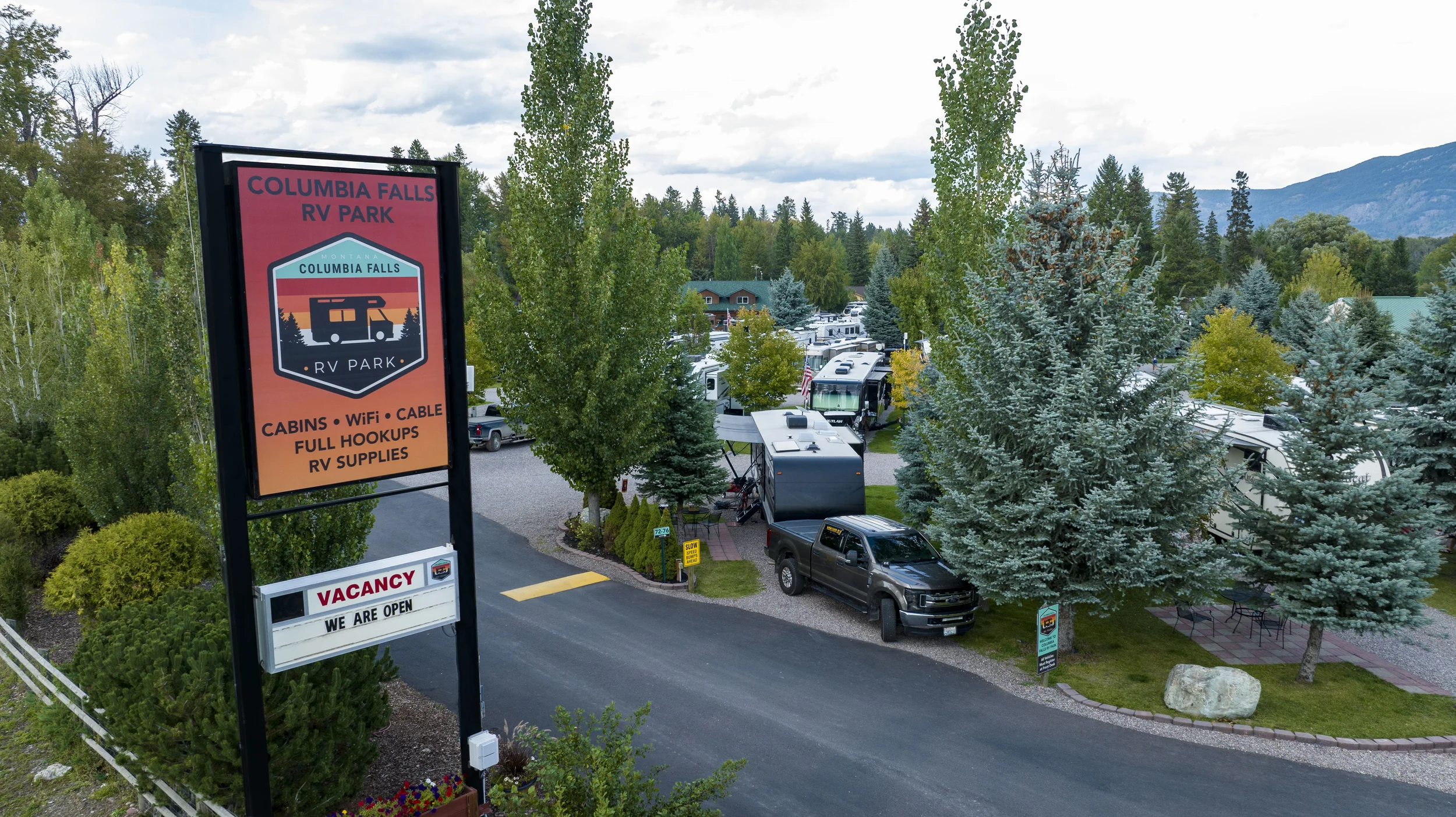 Columbia Falls RV Park entrance sign displaying amenities including cabins, WiFi, cable, full hookups, and RV supplies, with a vacancy notice, surrounded by mature pine and aspen trees with RVs parked